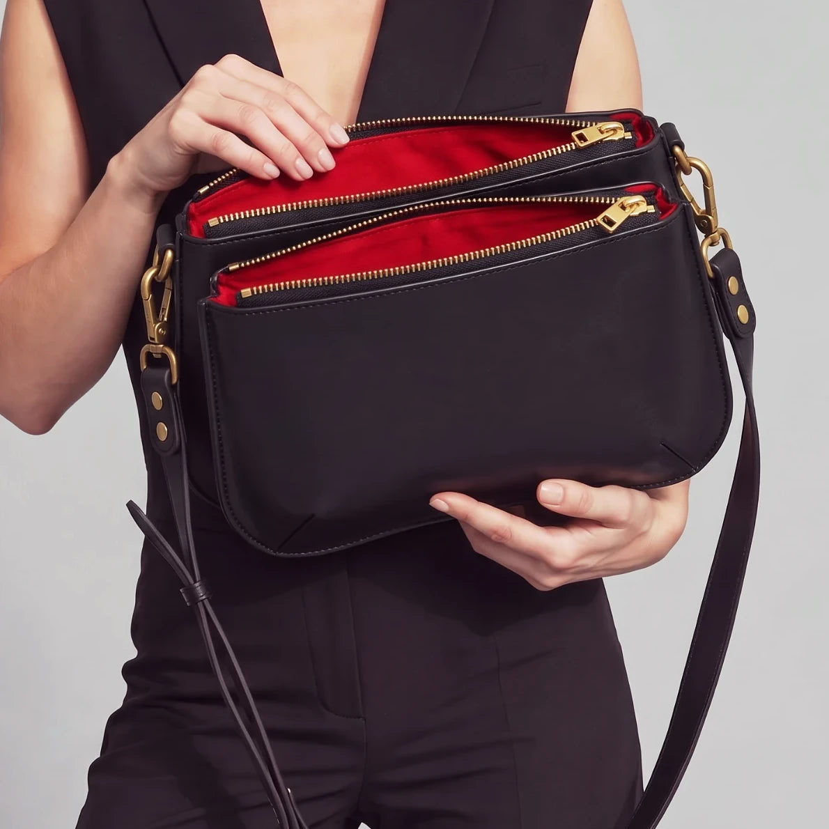 Deux mains présentent une base Baby Double en cuir noir, sans rabat, ses deux compartiments zippés ouverts sur la doublure rouge